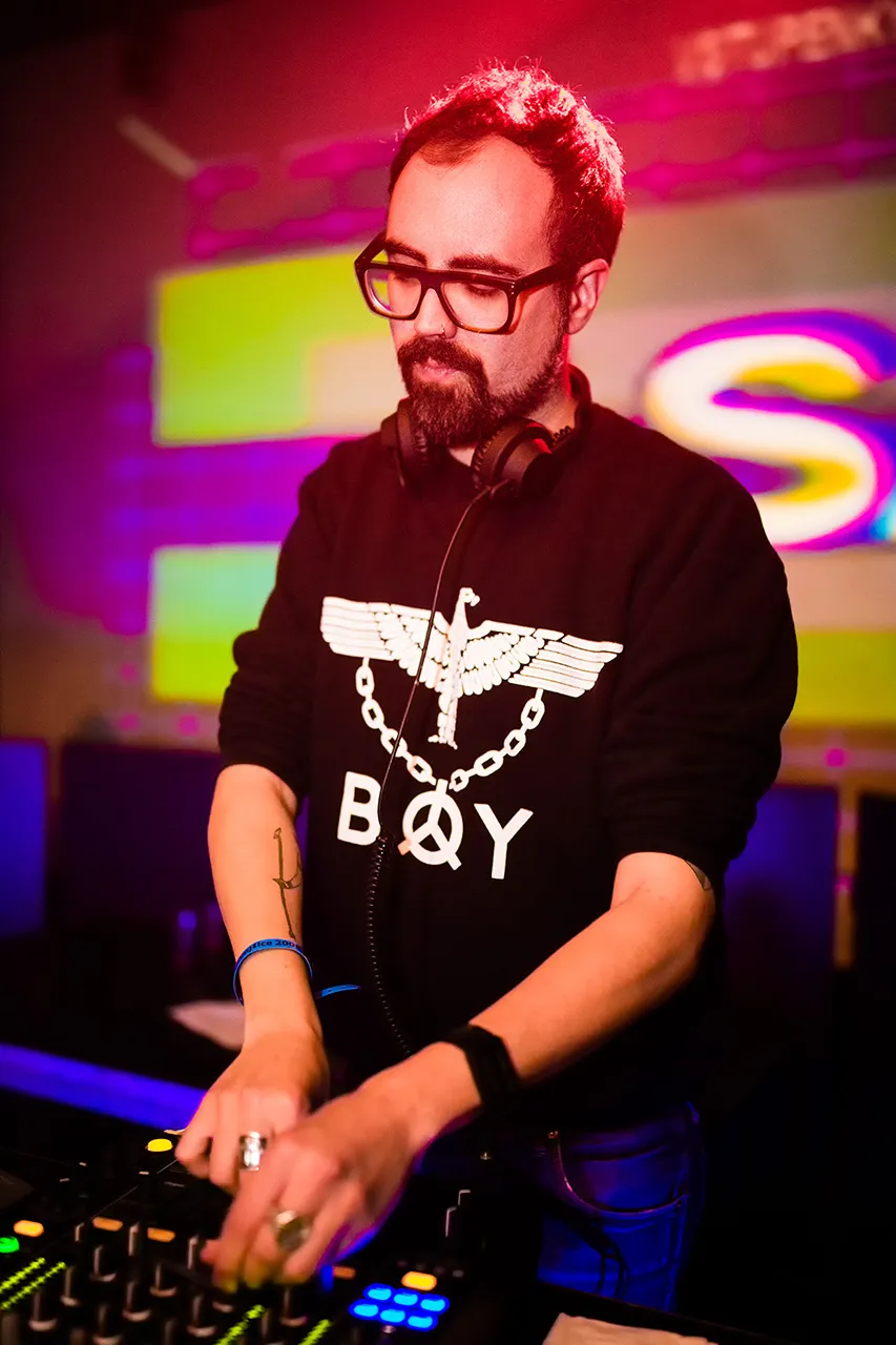 Muž s brýlemi a plnovousem, Henry Saiz, stojící za mixážním pultem, soustředící se na svůj mix v barevně nasvíceném klubu Roxy