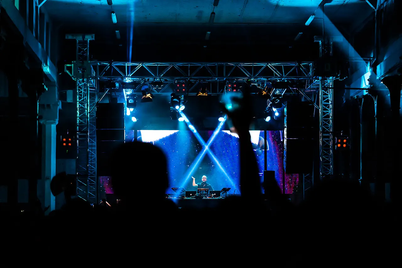 Pohled z publika na na stage barevně nasvícenou hudební stage s DJ, industriální prostředí halové taneční party