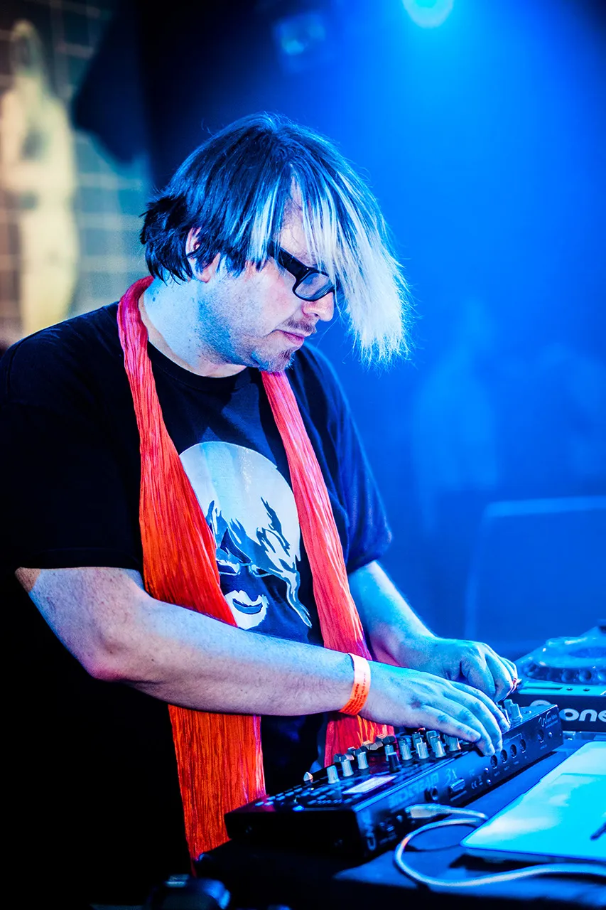 Muž s brýlemi, Robert Babicz, stojící za mixážním pultem, soustředící se na svůj mix v barevně nasvíceném klubu Roxy