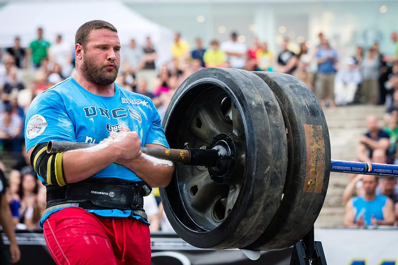 Závodník strongman v modrém tričku s rukama pokrytýma magnéziem svírá masivní železnou tyč se závažím v podobě velkých kol. Snímek zblízka zachycuje soustředění a fyzický tlak během disciplíny Conan's Wheel na závodech v Plzni.