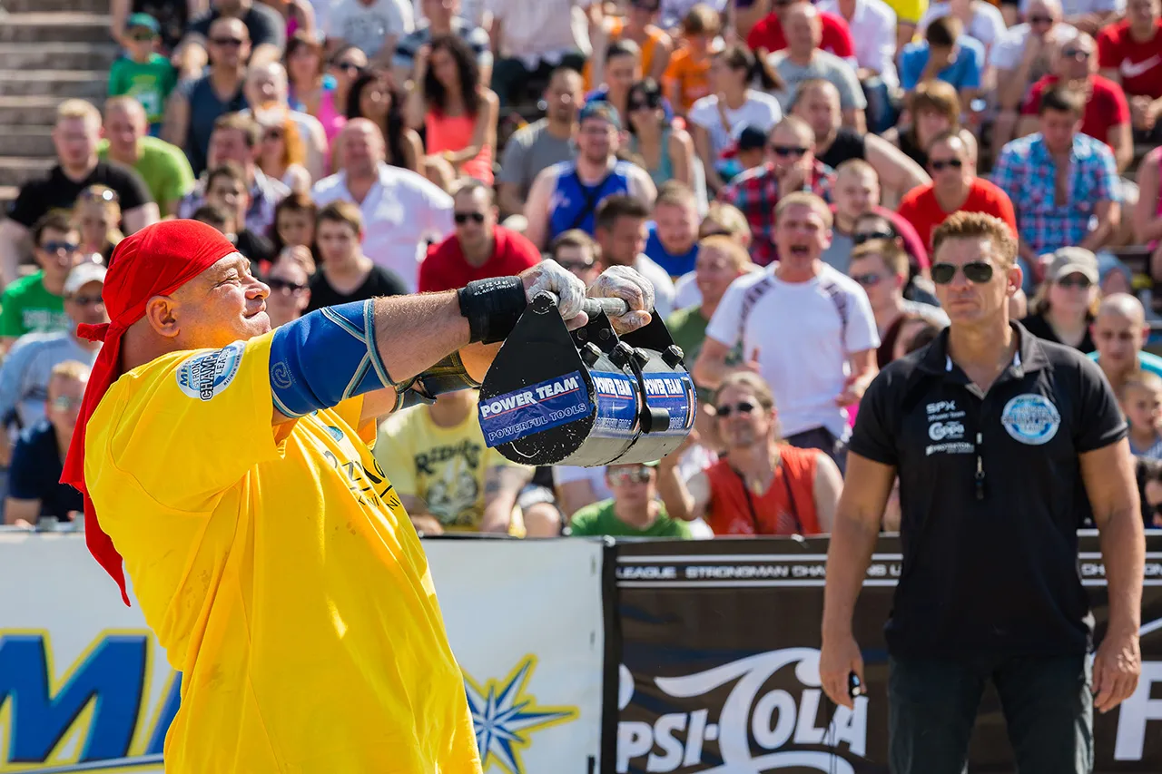 Strongman v červeném šátku drží v předpažení těžké válcové závaží během disciplíny Front Hold před zaplněnými tribunami.