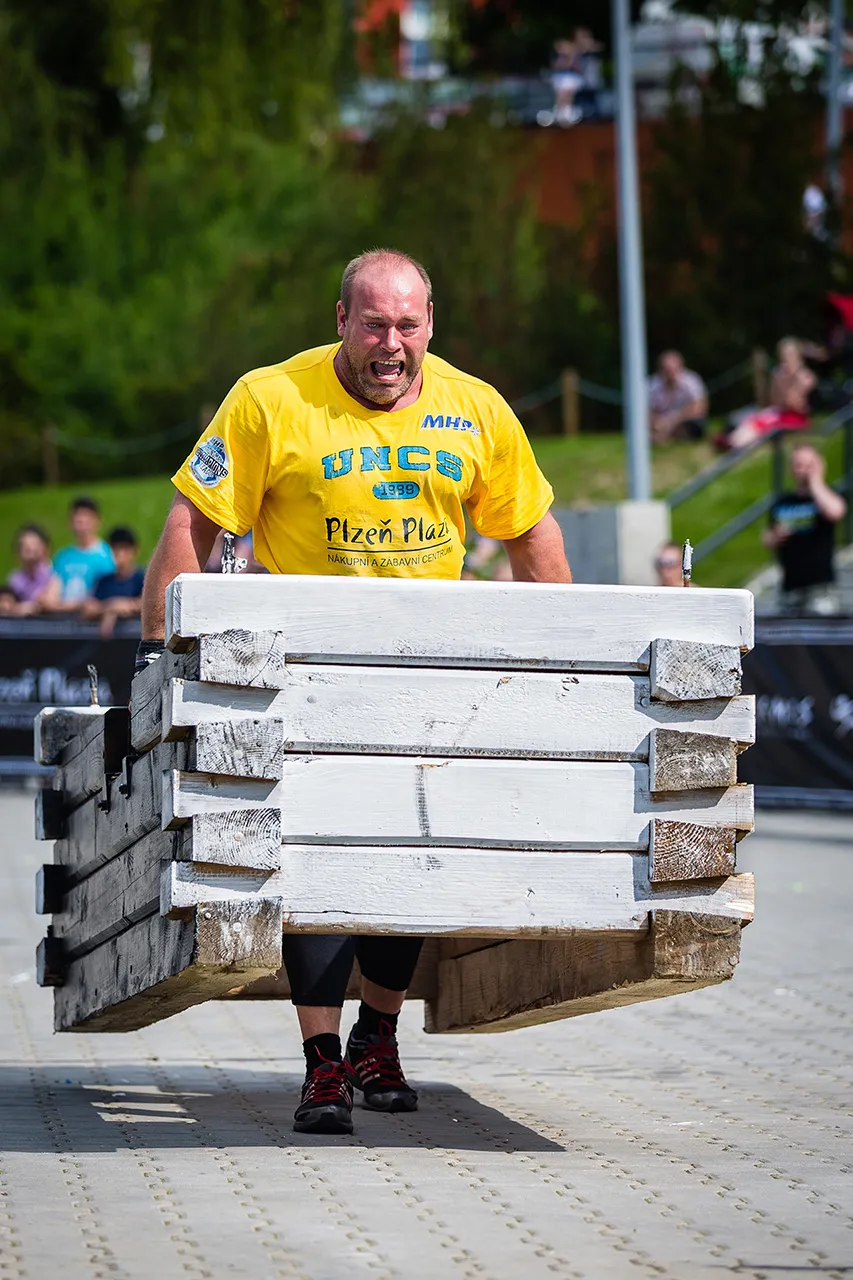 Strongman v plném nasazení při přenášení dřevěného rámu (Frame Carry). Fotografie z podhledu zdůrazňuje velikost břemene a odhodlání sportovce během závodu v Plzni.