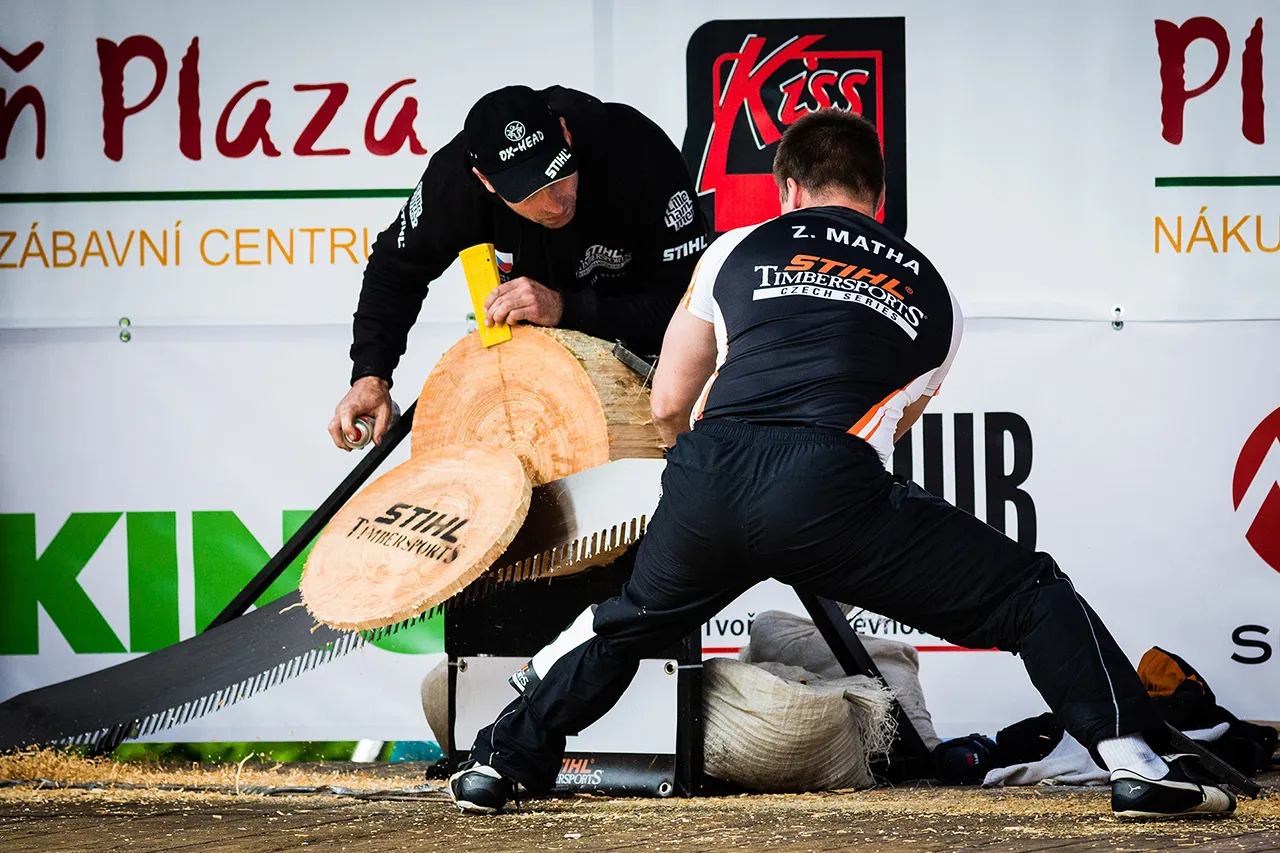 Pohled zezadu na závodníka Z. Mathu při disciplíně Single Buck na STIHL Timbersports. Na snímku je zachycen moment, kdy odřezaný kotouč s logem soutěže odpadává od klády, zatímco pomocník promazává list pily pro hladší řez.