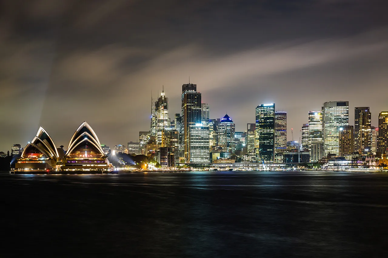 Noční panorama Sydney s osvětlenou budovou Opery a mrakodrapy v pozadí pod mírně zataženou oblohou.