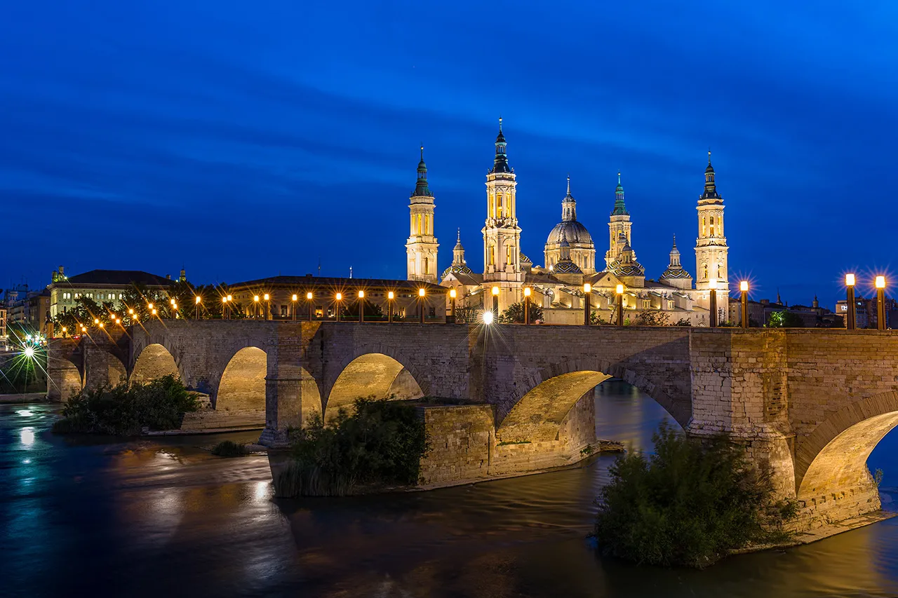Noční pohled na osvětlenou baziliku Panny Marie v Zaragoze přes kamenný most Puente de Piedra.
